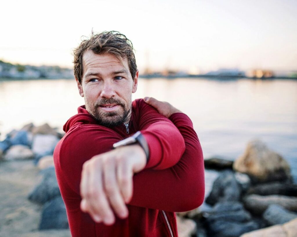 Keski-ikäinen mies venyttelee rannalla punaisessa hupparissa ja urheilukello kädessä. Miesten vaihdevuodet ja niiden oireet voivat helpottaa liikunnalla.