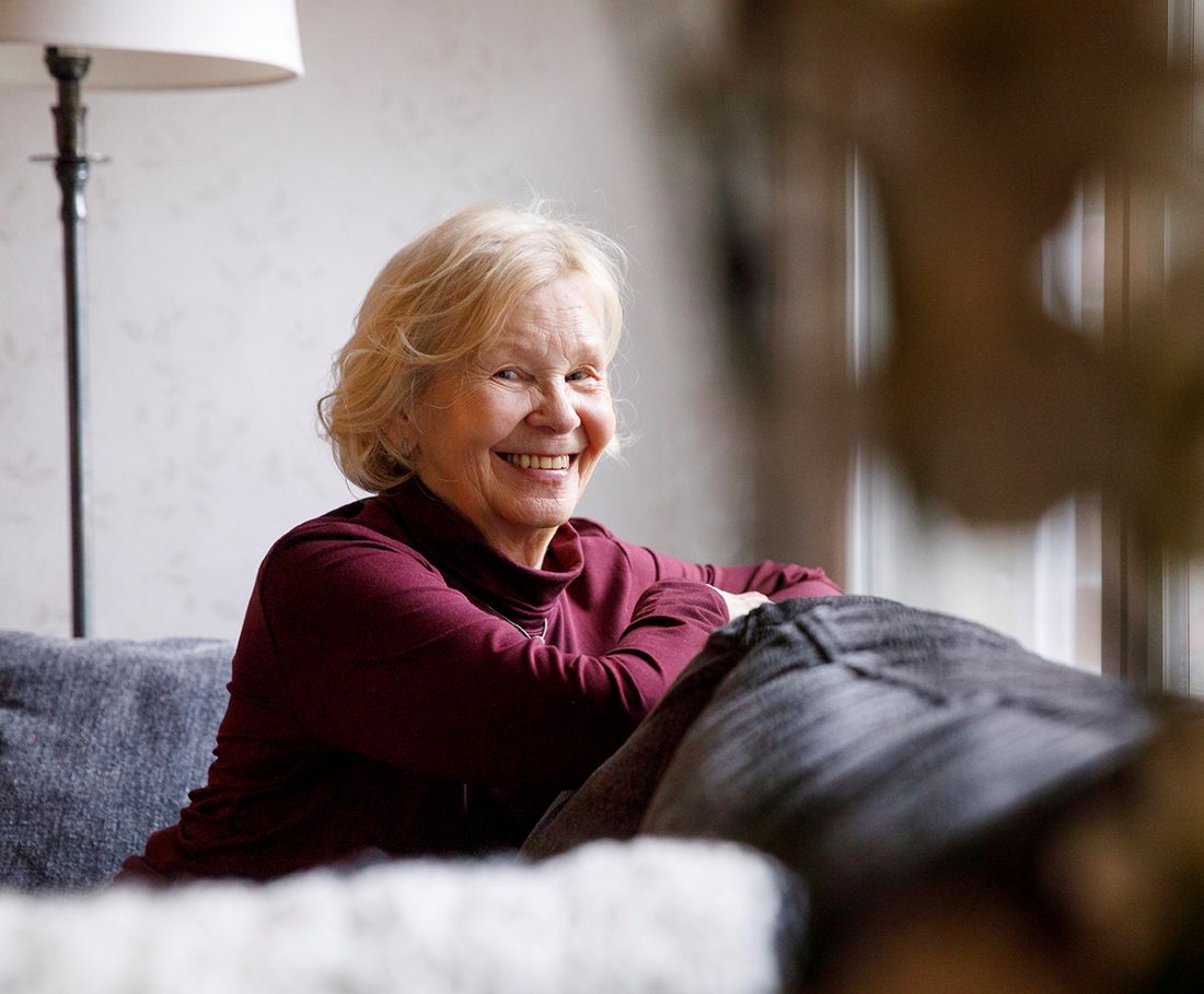 Kuvassa sohvalla istuu ja hymyilee iäkkäämpi nainen, jolla on luomireunan tulehdus.